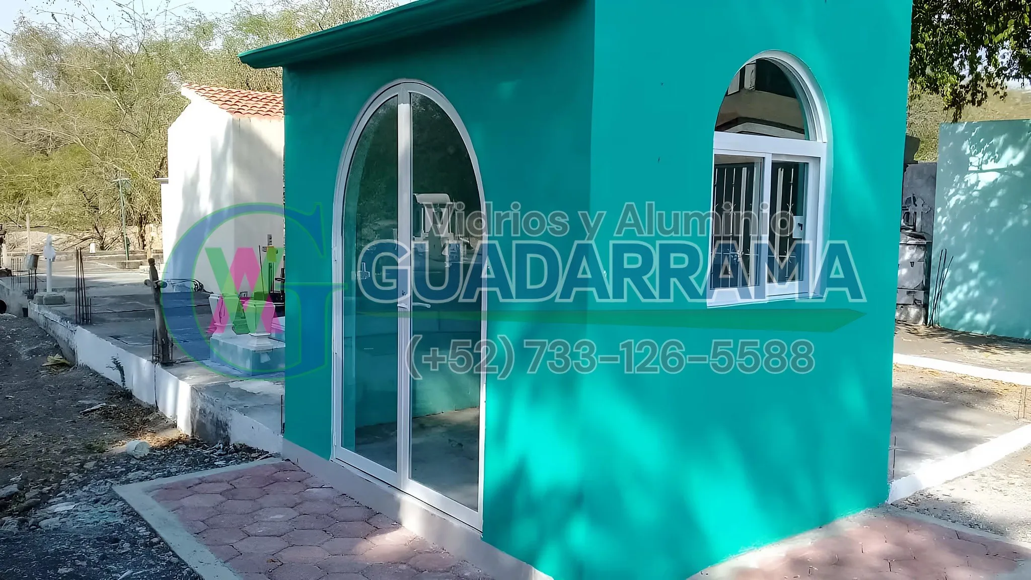 Puerta abatible con arco de medio punto en aluminio blanco y vidrio templado transparente, instalada en una construcción reciente en tonos turquesa.